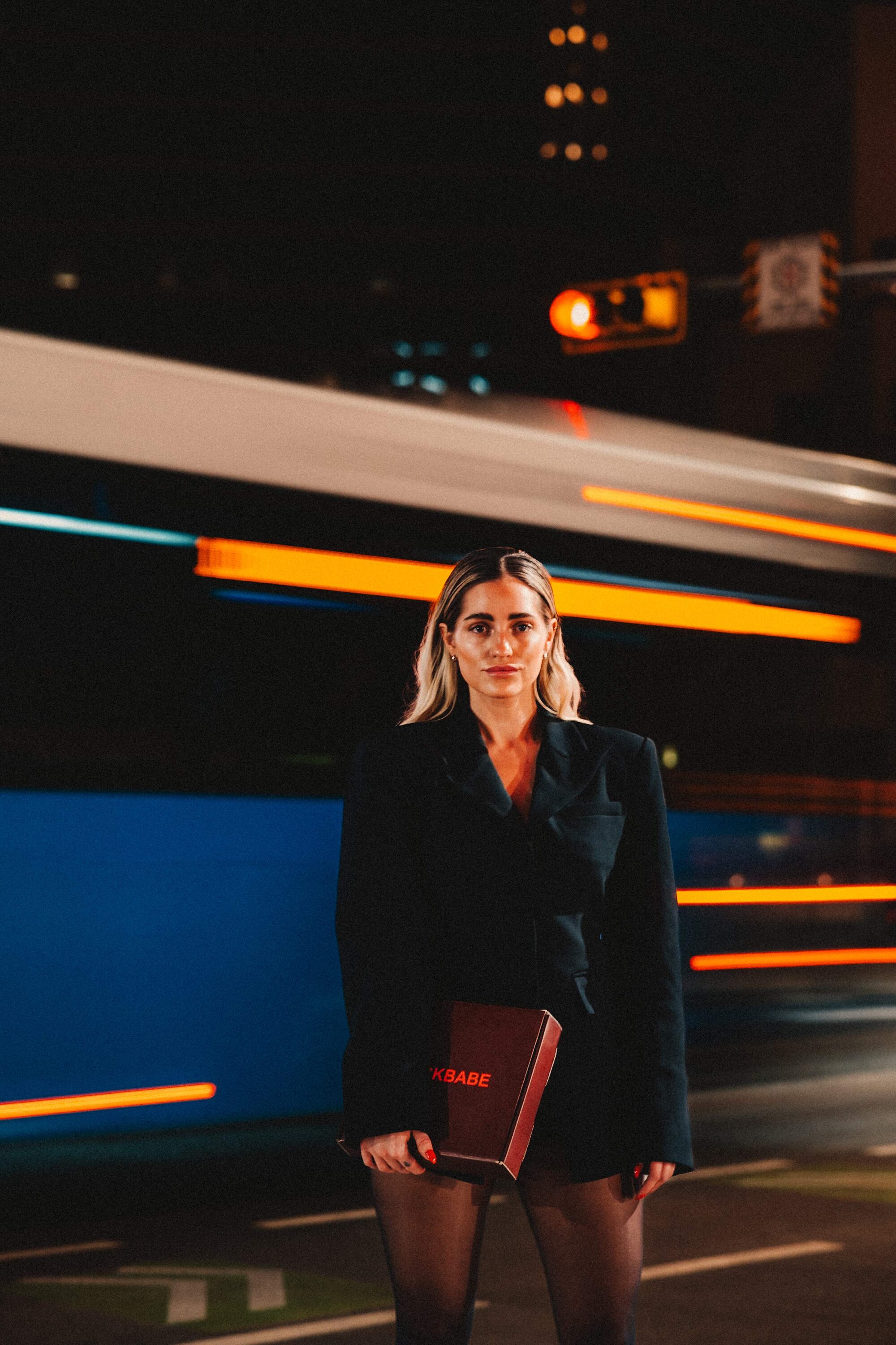 Casey holding SLICKBABE box on urban night street with light trails — black blazer, red nails, editorial campaign shot